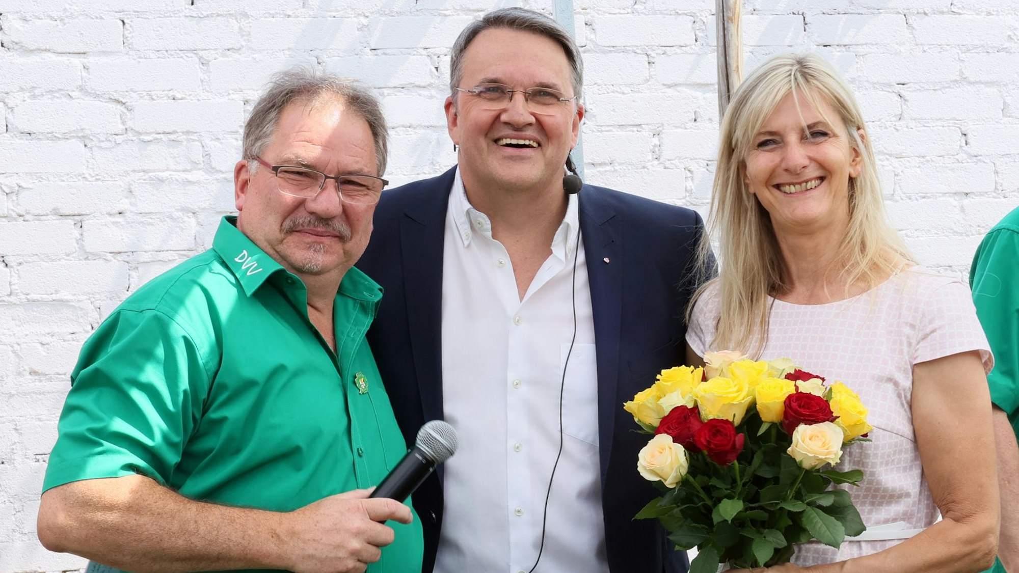 Links steht Andreas Molter mit grünem Hemd. Er hält ein Mikrofon in der Hand. In der Mitte steht Udo Becker im Anzug. Rechts steht die Frau von Udo Becker mit einem Blumenstrauß in der Hand.