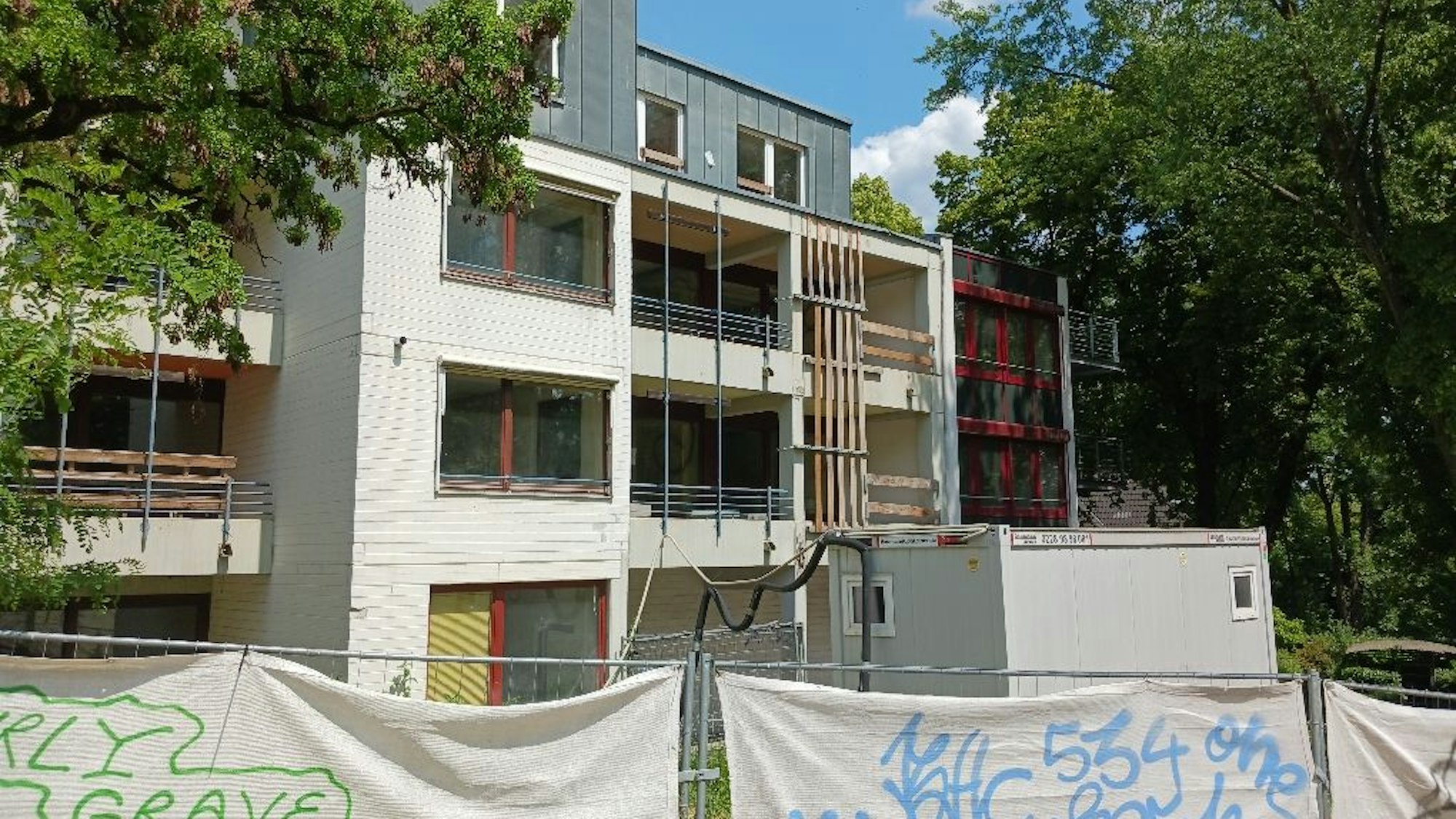 Ein Bauzaun steht vor einem mehrstöckigen Gebäude, das von Bäumen gesäumt wird.