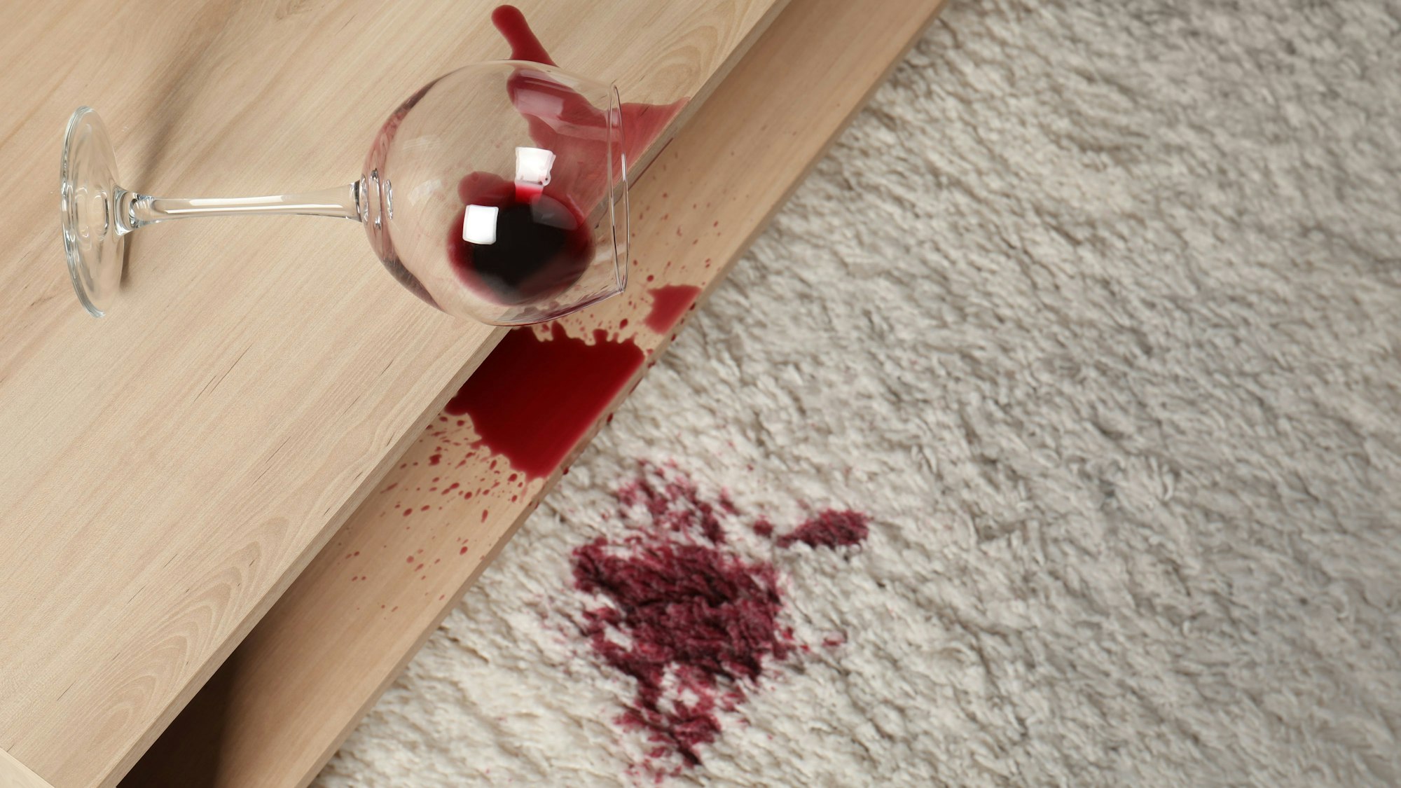 Ein verschüttetes Glas Rotwein auf weißem Teppich.