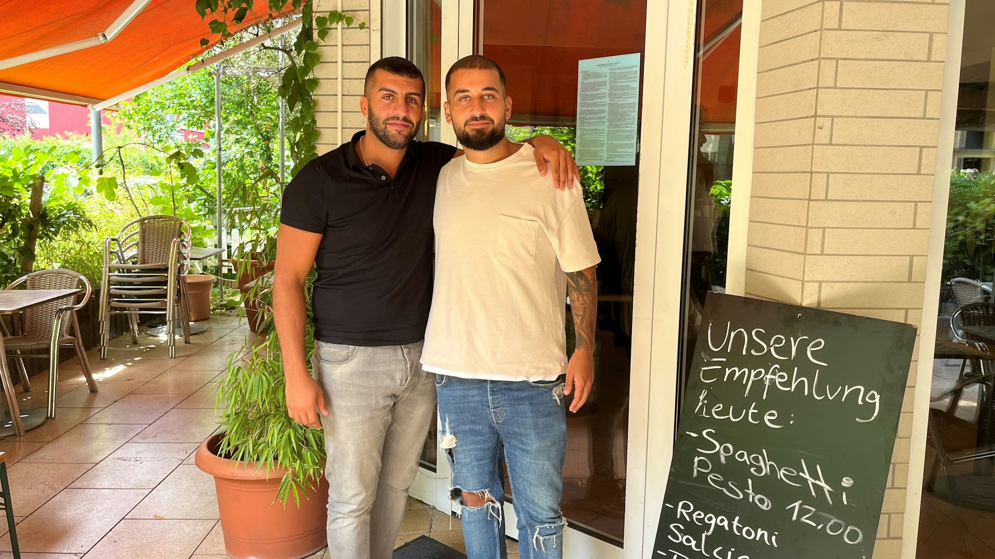 Zwei junge Männer mit dunklen Haaren stehen nebeneinander auf einer Terrasse vor einem Restaurant-Eingang.