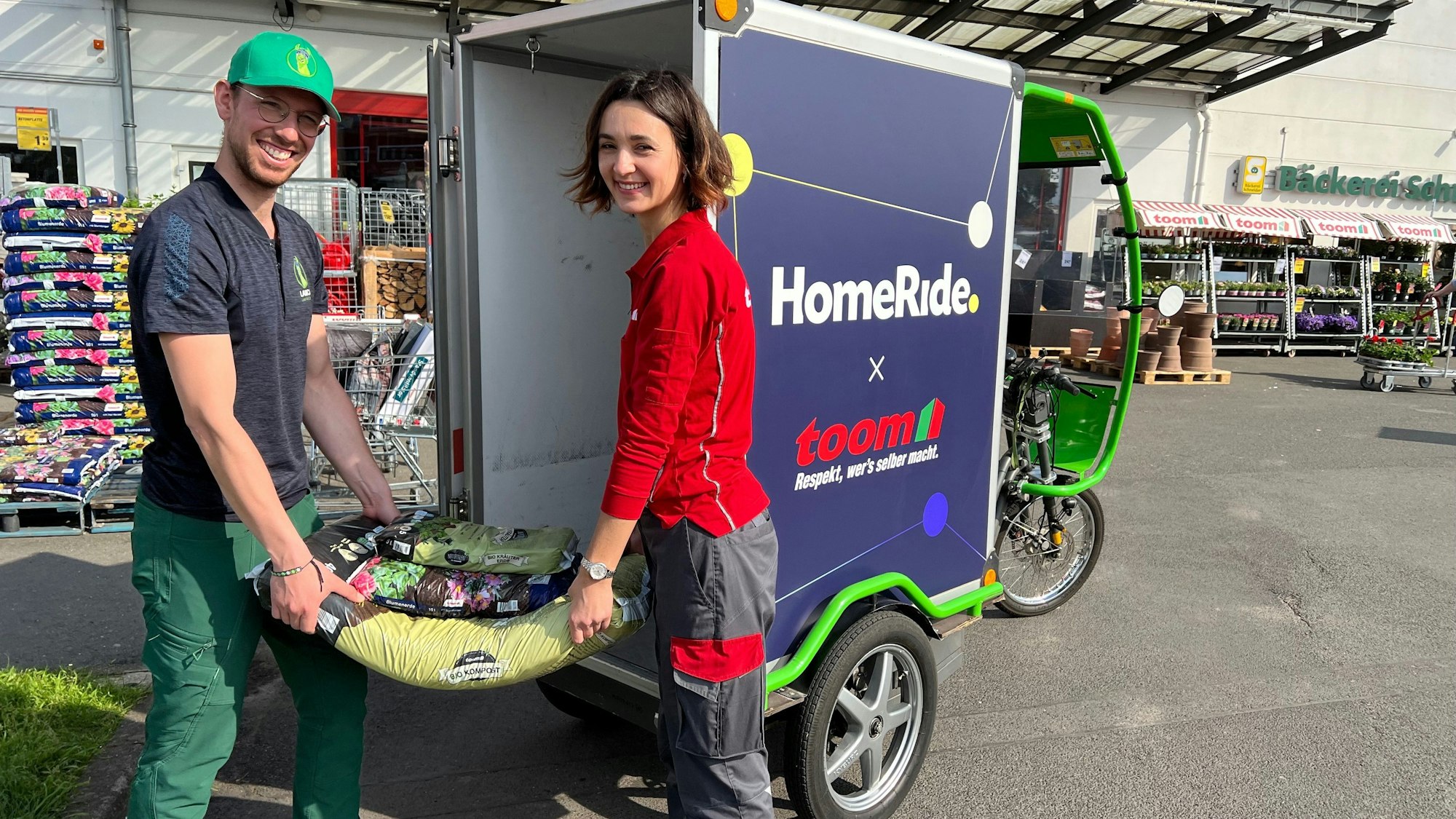 Mitarbeitende der Kölner Liefer-App Homeride verladen Blumenerde vor einem Baumarkt in ein Lastenfahrrad.