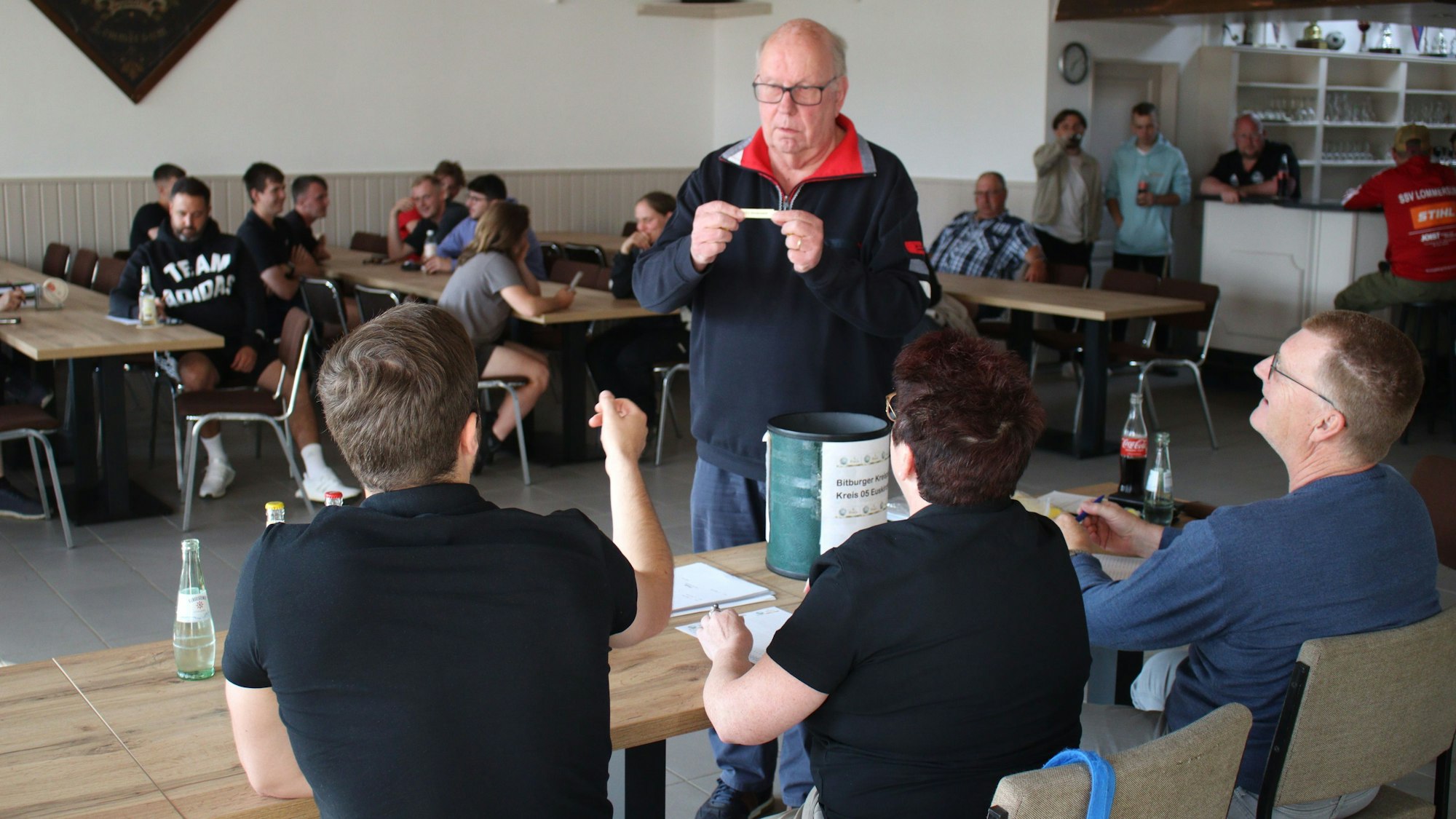 Heinrich van Wersch zeigt den Verantwortlichen des Fußballkreises eines der gezogenen Lose. Im Hintergrund sitzen Vertreter einiger Vereine.