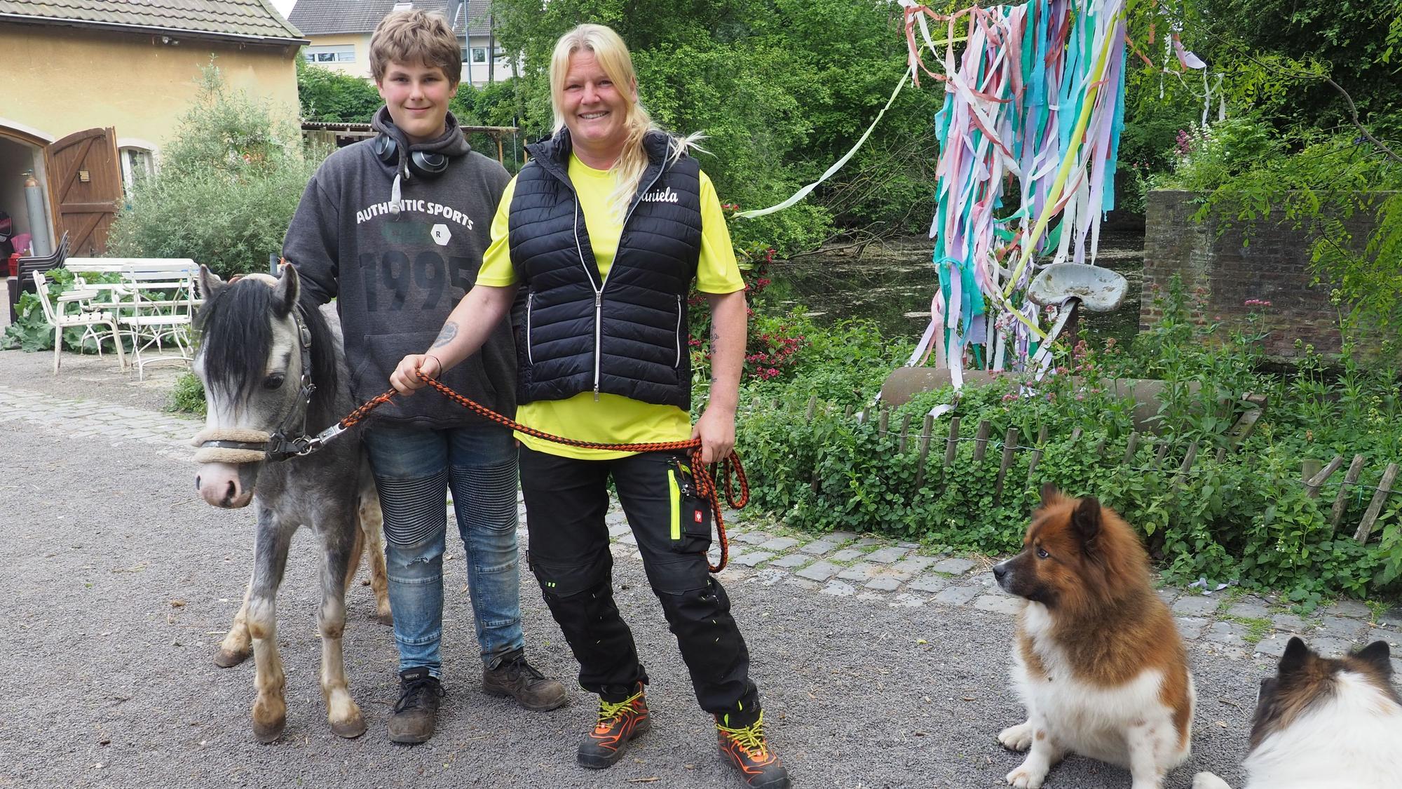Daniela Schmitz steht nebem einem Jungen, Maik Schneider, der ein graues Pony streichelt. Zwei kleine Hunde liegen neben ihnen.