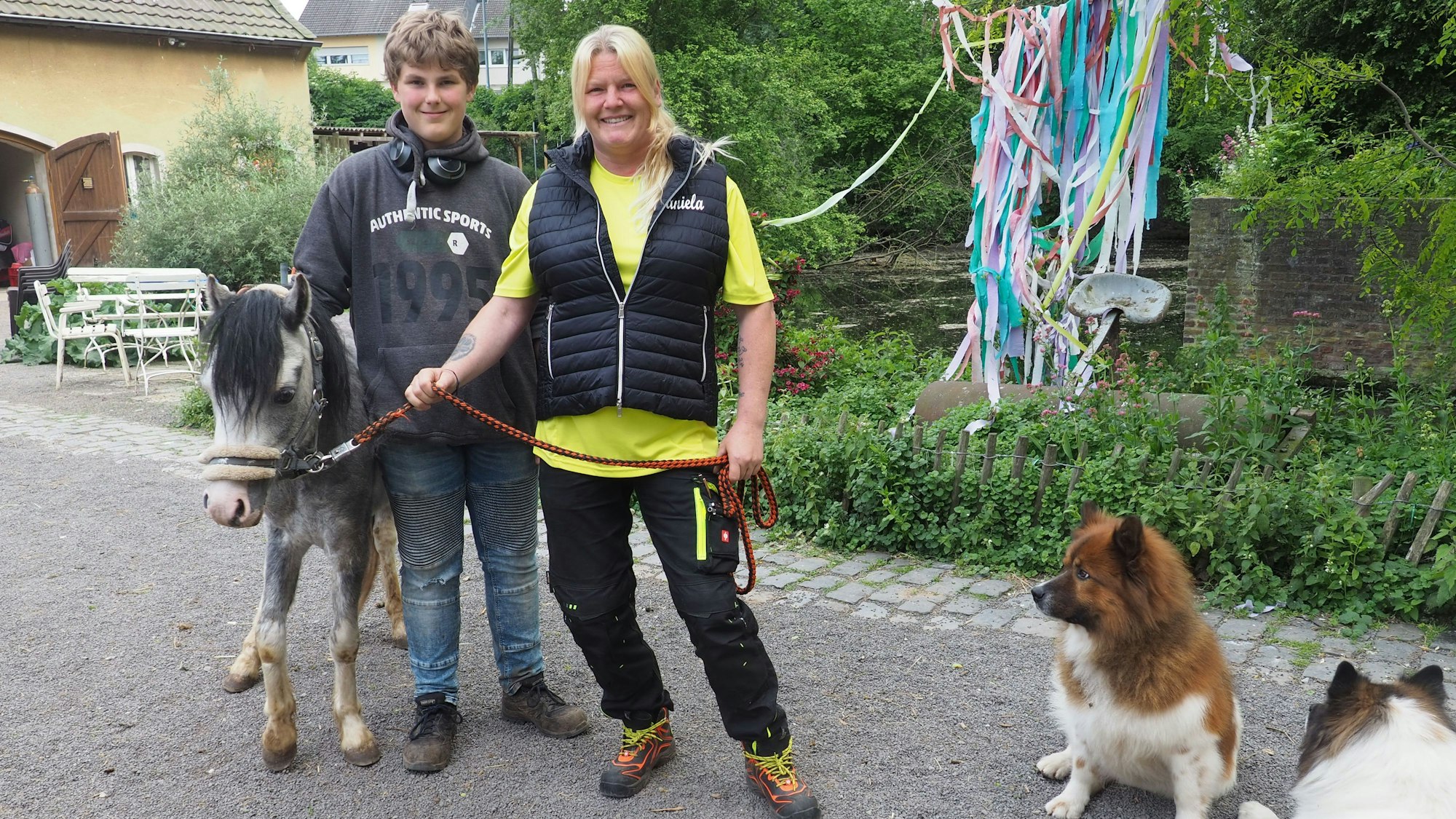 Daniela Schmitz steht nebem einem Jungen, Maik Schneider, der ein graues Pony streichelt. Zwei kleine Hunde liegen neben ihnen.