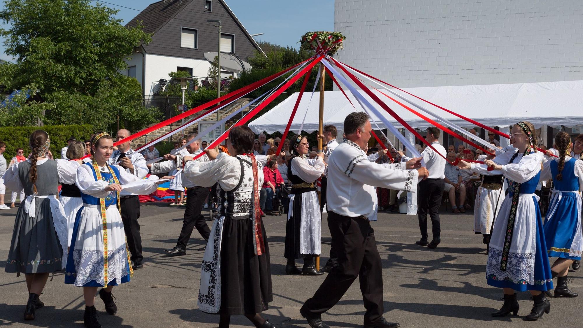 Männer und Frauen in bunten Trachten tanzen mit Bändern im Kreis.