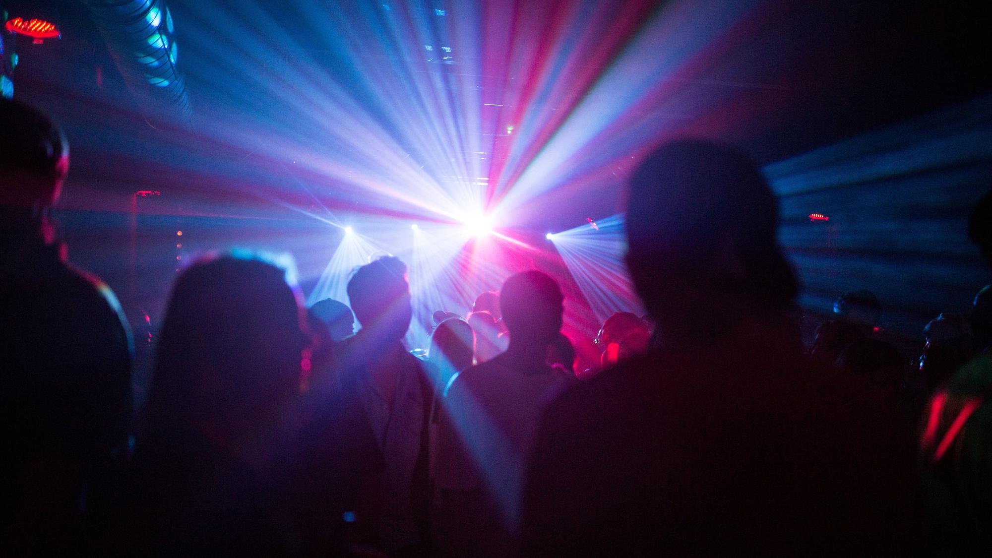 Eine Gruppe von Menschen steht mit dem Rücken zur Kamera in einem Club und wird von Lichtstrahlern angeleuchtet.