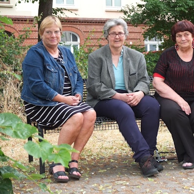 Drei ältere Damen sitzen auf einer Bank, eine stützt den Arm auf ihren Rollator.