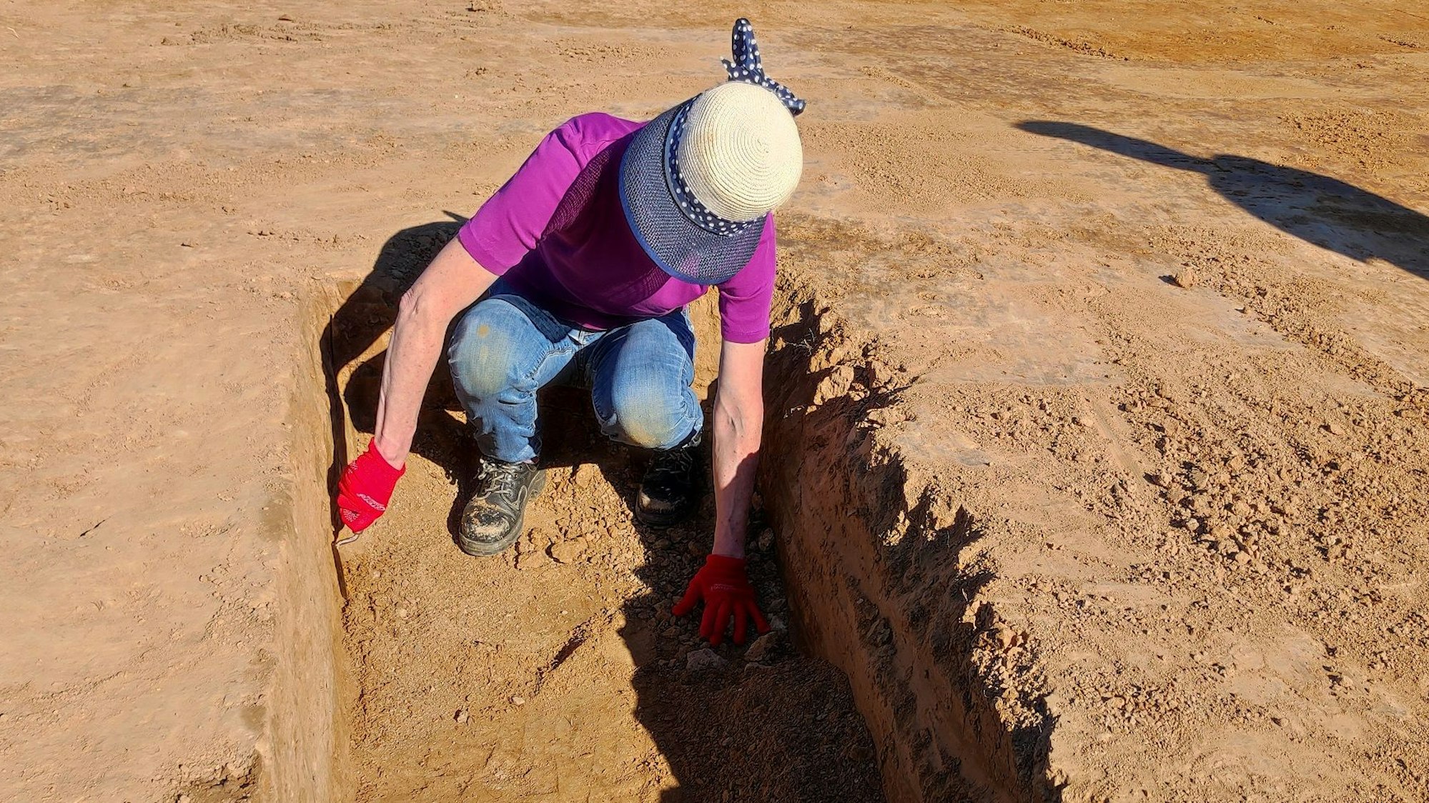 Das Bild zeigt eine Grabungshelferin, die mit einer Keller sandigen Boden zur Seite schiebt.