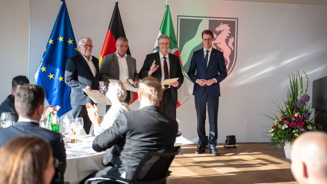 Wolf-Christian Lorenz, Carsten Bönsch, Hubert Schilles und Ministerpräsident Hendrik Wüst stehen in der Staatskanzlei vor einem den Flaggen von der Europäischen Union, Deutschland und Nordrhein-Westfalen. Sie werden von einem sitzenden Publikum beklatscht.
