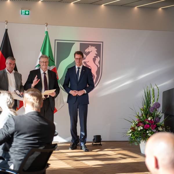 Wolf-Christian Lorenz, Carsten Bönsch, Hubert Schilles und Ministerpräsident Hendrik Wüst stehen in der Staatskanzlei vor einem den Flaggen von der Europäischen Union, Deutschland und Nordrhein-Westfalen. Sie werden von einem sitzenden Publikum beklatscht.