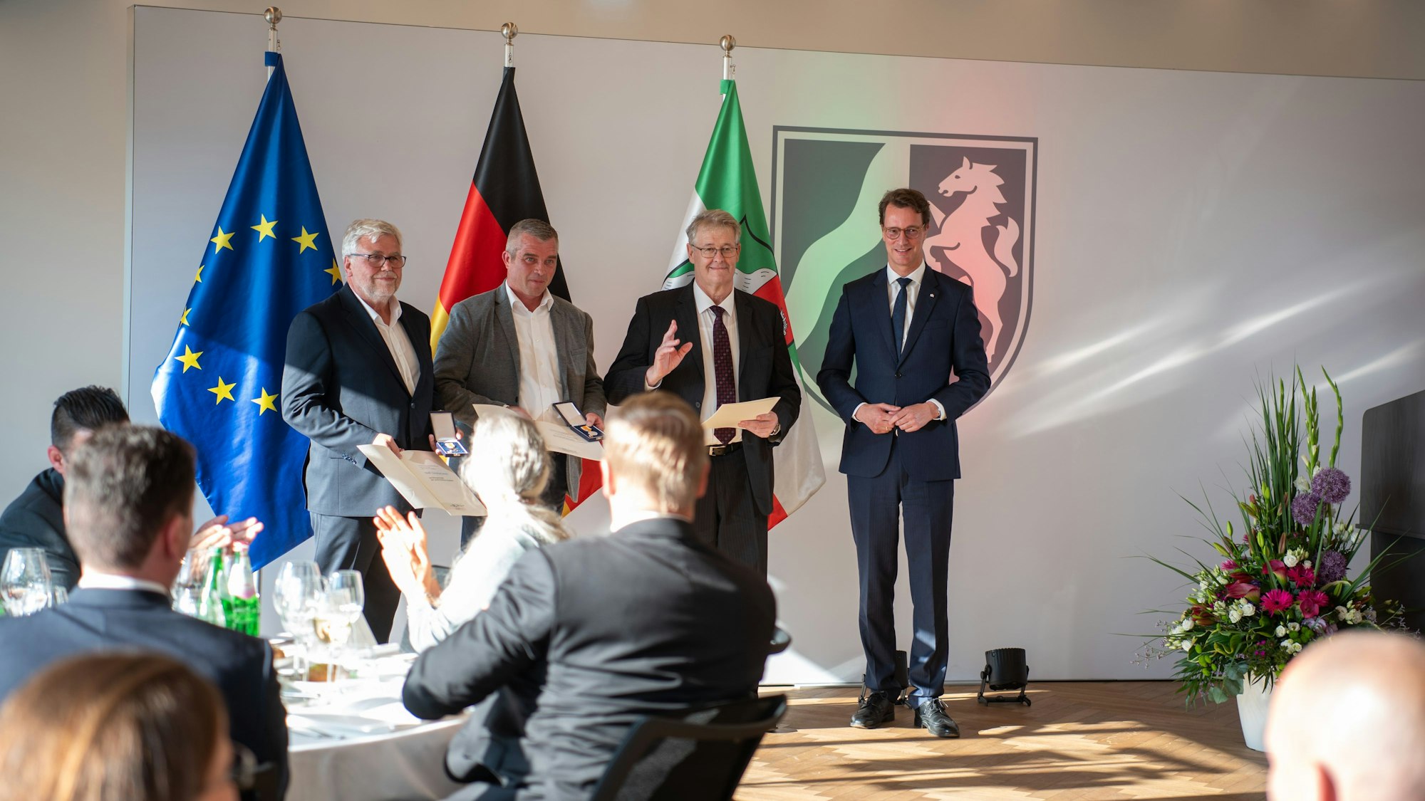 Wolf-Christian Lorenz, Carsten Bönsch, Hubert Schilles und Ministerpräsident Hendrik Wüst stehen in der Staatskanzlei vor einem den Flaggen von der Europäischen Union, Deutschland und Nordrhein-Westfalen. Sie werden von einem sitzenden Publikum beklatscht.