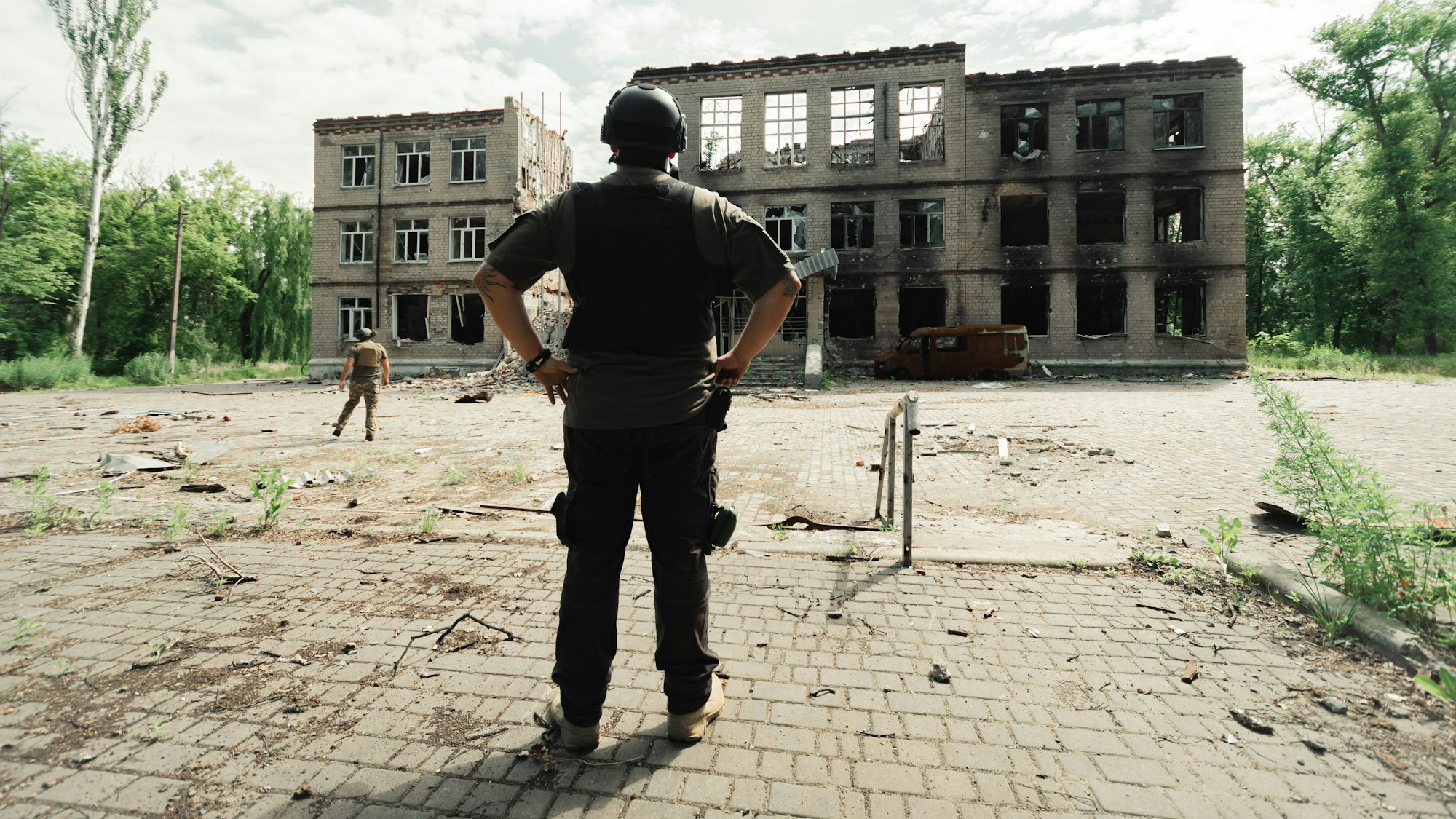 Ein Mann mit Helm und schusssicherer Weste steht vor einem zerbombten Gebäude.