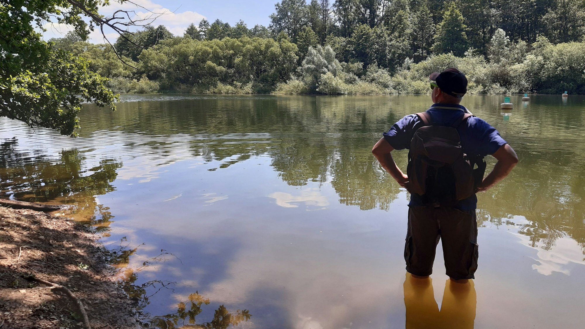 Ein Wanderer steht bis zu den Knien im Wasser am Ufer eines Stausees.