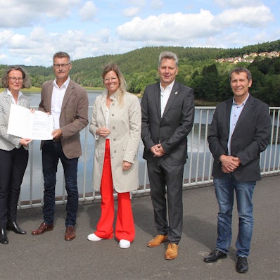 Die Ministerin überreicht Bürgermeister Lembach auf dem Damm des Kronenburger Sees den Bescheid, daneben die Landräte und kommunale Vertreter.