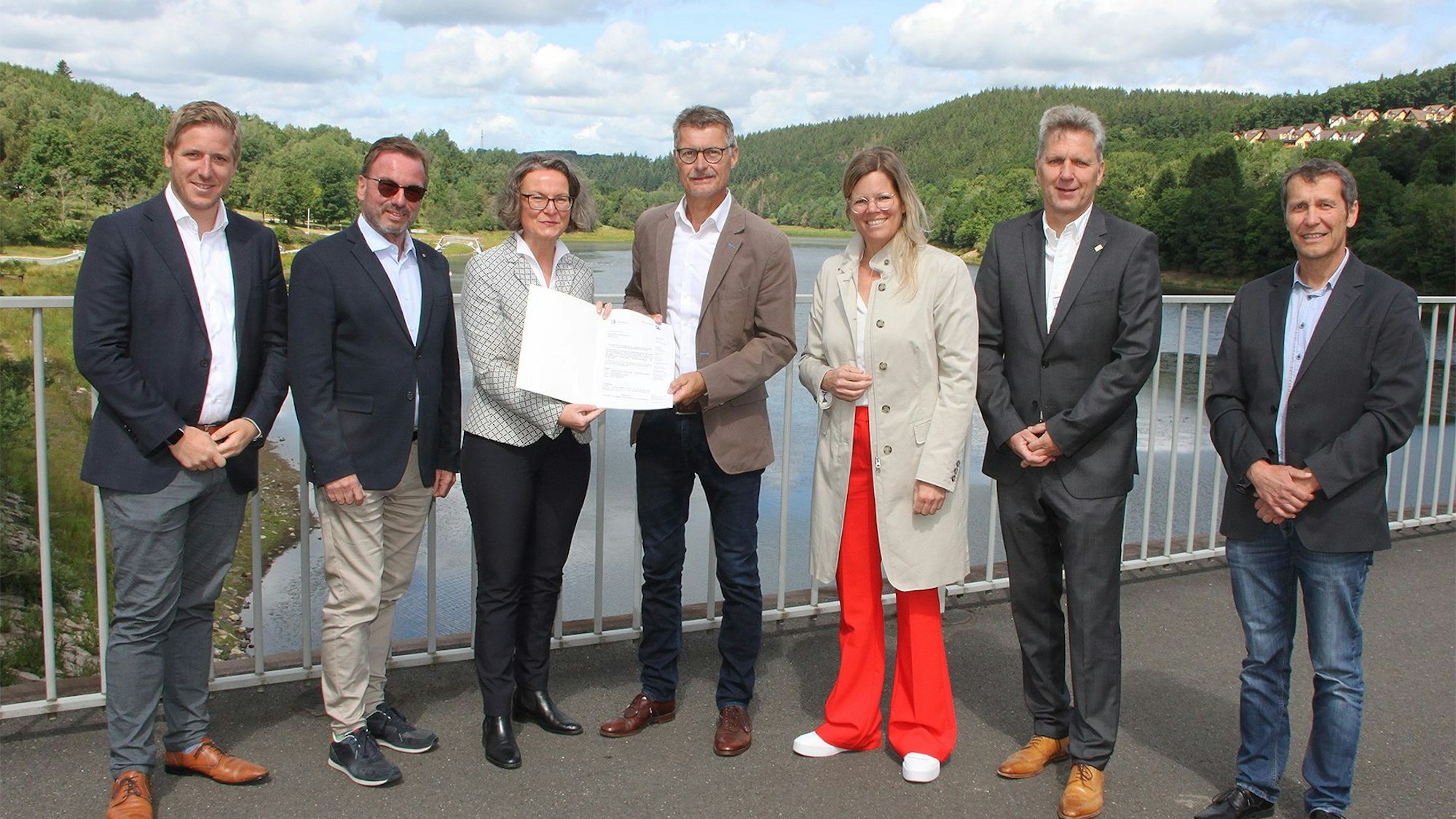 Die Ministerin überreicht Bürgermeister Lembach auf dem Damm des Kronenburger Sees den Bescheid, daneben die Landräte und kommunale Vertreter.