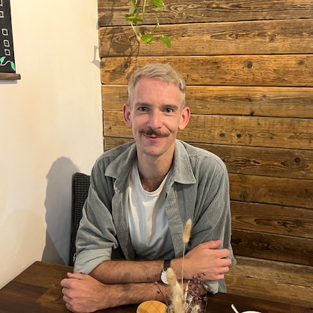 Ein junger Mann mit Schnurrbart sitzt an einem Tisch, mit den Armen über kreuz aufgestützt.