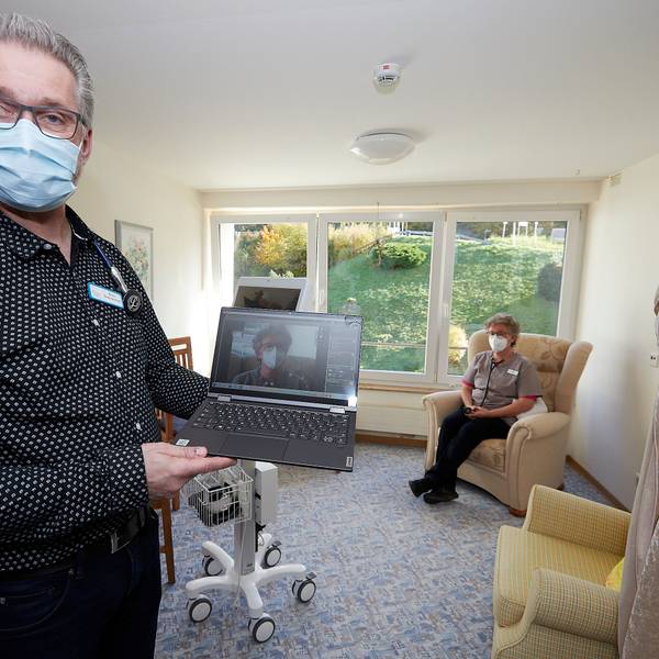 Pflegedienstleiter Arno Brauckmann hält ein Notebook in der Hand und demonstriert, was ein Arzt bei der Telemedizin sehen würde. Im Hintergrund sitzt eine Patientin.