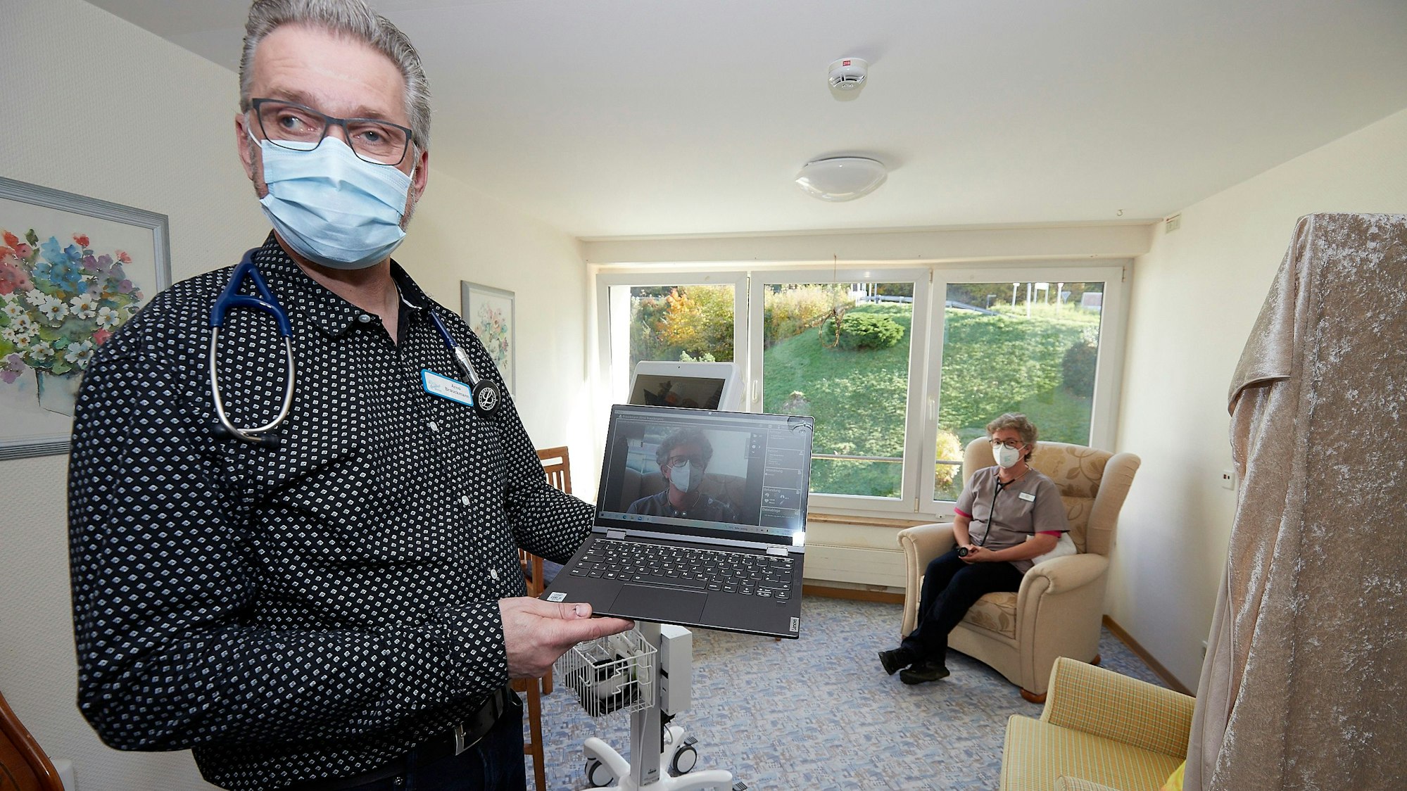 Pflegedienstleiter Arno Brauckmann hält ein Notebook in der Hand und demonstriert, was ein Arzt bei der Telemedizin sehen würde. Im Hintergrund sitzt eine Patientin.