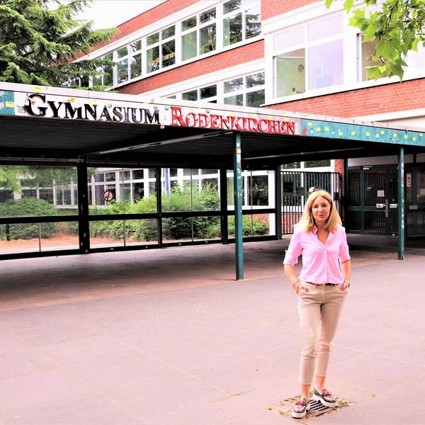 Eine blonde Frau mit schulterlangem blonden Haar steht vor einem Gebäude, auf dem der Schriftzug Gymnasium Rodenkirchen zu lesen ist.
