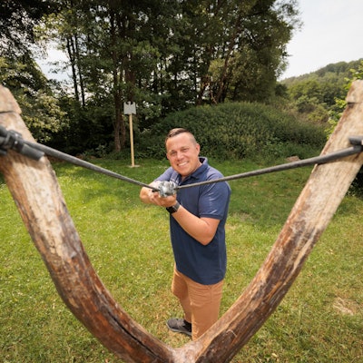 Ein Mann spannt eine Riesenzwille und zielt lächelnd auf den Fotografen.