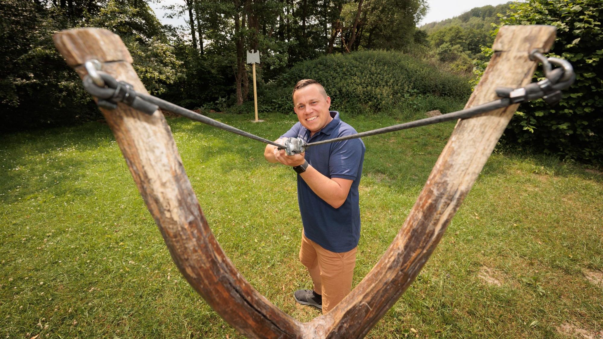 Ein Mann spannt eine Riesenzwille und zielt lächelnd auf den Fotografen.