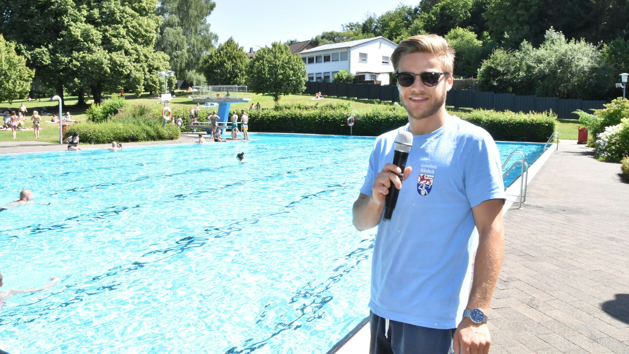 Am Becken eines Freibads steht ein junger Mann mit einem Mikrofon, das ersetzt der Badeaufsicht die Trillerpfeife.