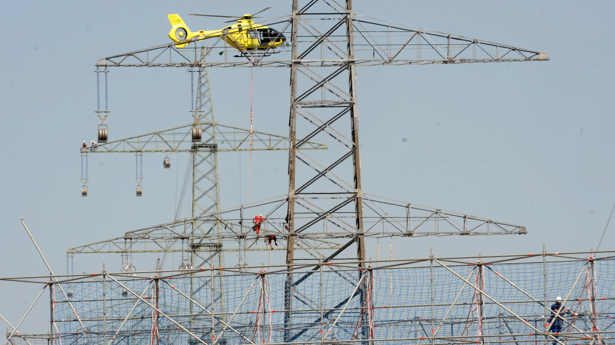 Das Bild zeigt Monteure auf einem Hochspannungsmast und einen Hubschrauber, der eine Leitung an einem Seil dort hinbringt.