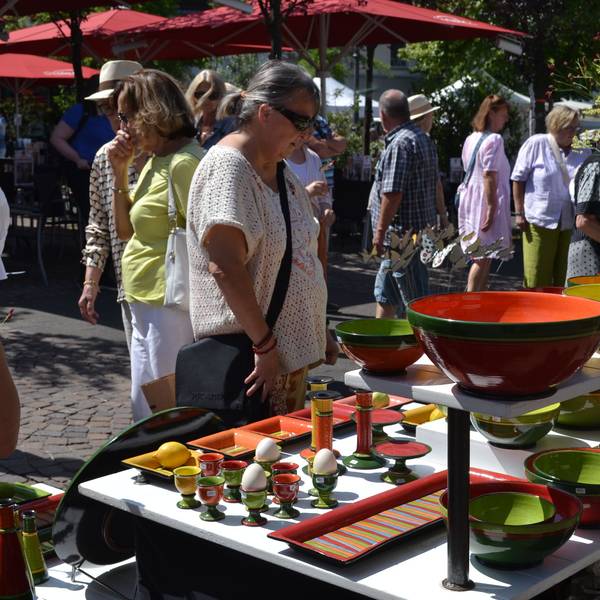 Bunte Vasen, Schüsseln und Eierbecher stehen auf einem mehrstöckigen Ausstellungstisch, an dem Besucher im Sonnenschein stehen.