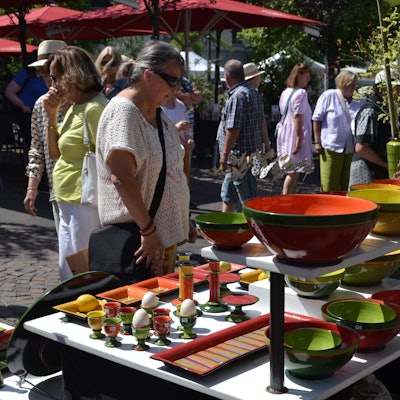 Bunte Vasen, Schüsseln und Eierbecher stehen auf einem mehrstöckigen Ausstellungstisch, an dem Besucher im Sonnenschein stehen.