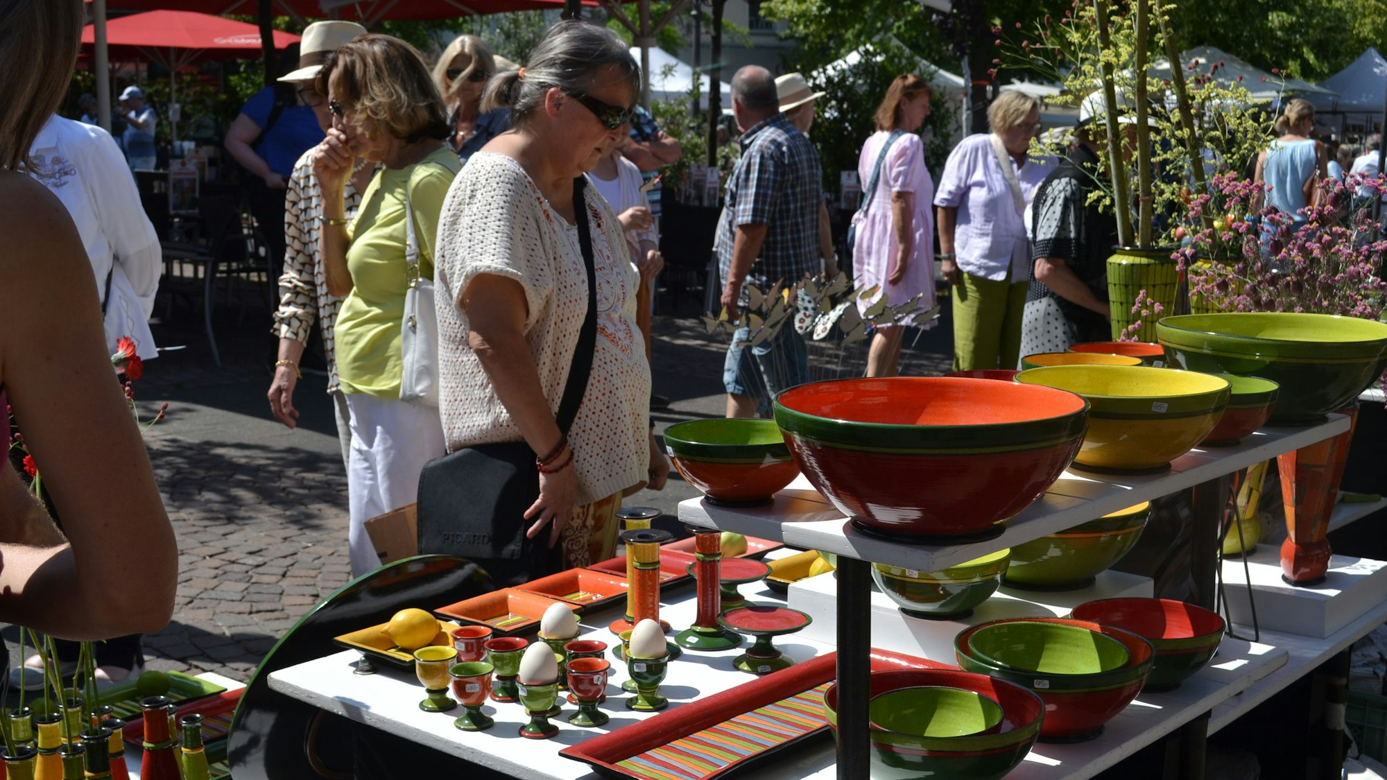 Bunte Vasen, Schüsseln und Eierbecher stehen auf einem mehrstöckigen Ausstellungstisch, an dem Besucher im Sonnenschein stehen.