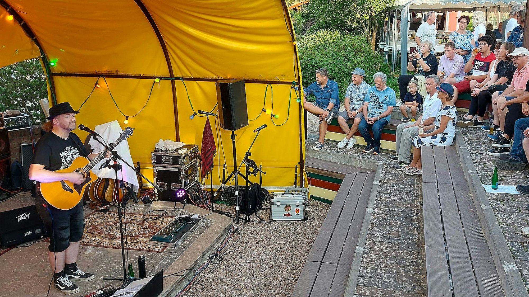 Ein Mann mit schwarzem Hut steht links an einem Mikrofon, singt und spielt Gitarre. Auf einer Tribüne vor ihm sitzen einige Menschen, sie tragen Sommerkleidung.