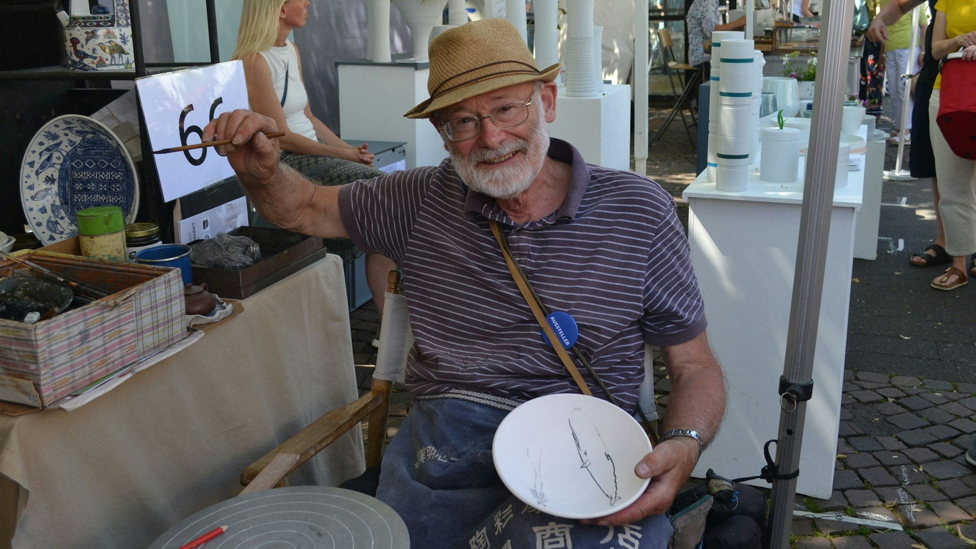 Ein älterer Mann mit Strohhut hat die Hand mit einem feinen Pinsel erhoben. In der anderen Hand hält er eine weiße Schüssel, auf die er bereits dünne Linien malte.