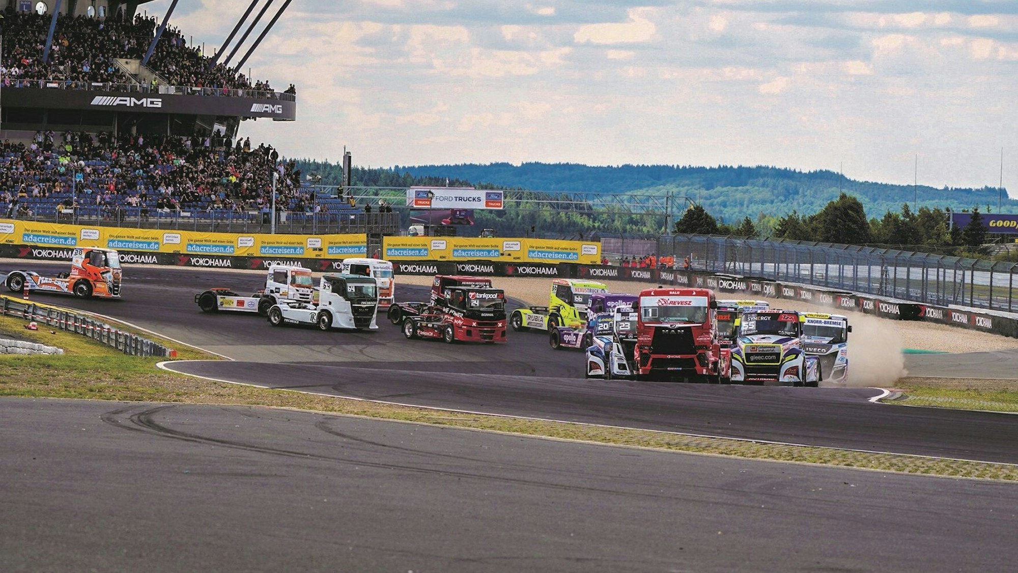 Mehrere Trucks liefern sich ein Rennen. Im Hintergrund sind eine Tribüne mit vielen Zuschauern und Wald zu erkennen.