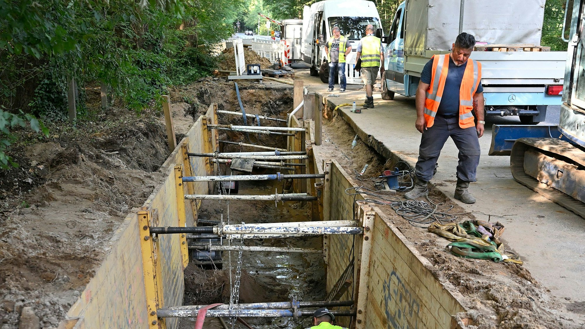 Blick in die Tiefe einer Kanalbaustelle am Straßenrand. Bauarbeiter und ihre Fahrzeuge sind zu sehen.