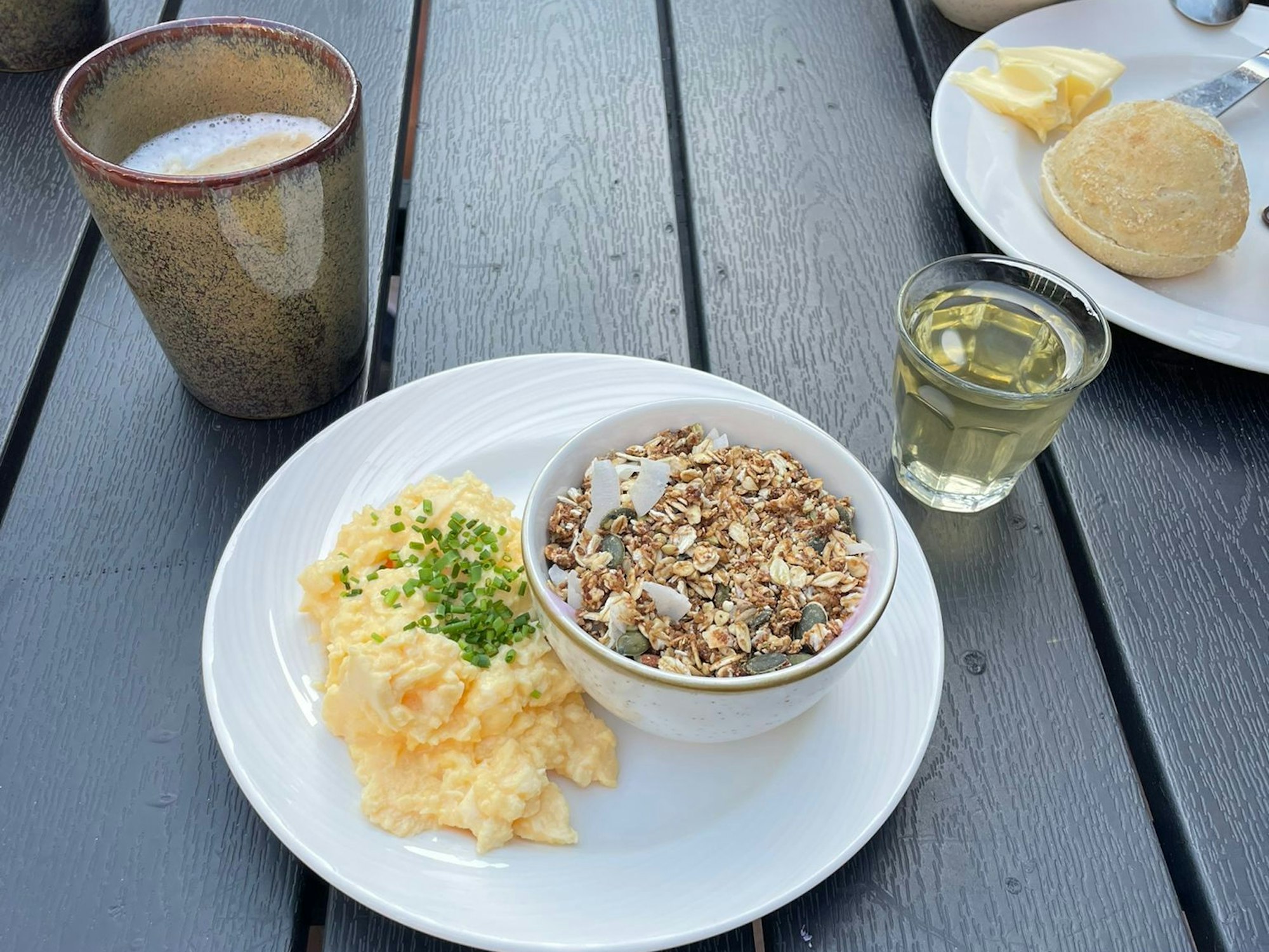 Frühstück: Auf einem Teller stehen eine gefüllte Müsli-Schale und Rührei, dahinter stehen ein Caffe Latte und ein Glas Apfelsaft. Im Hintergrund ein Teller mit einem Brötchen.
