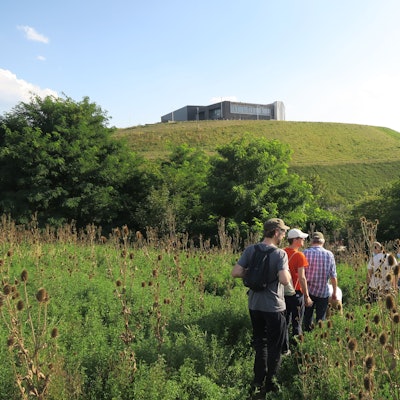 Menschen laufen durch ein Feld, im Hintergrund ist ein Hügel mit einem Flachbau obendrauf zu sehen.