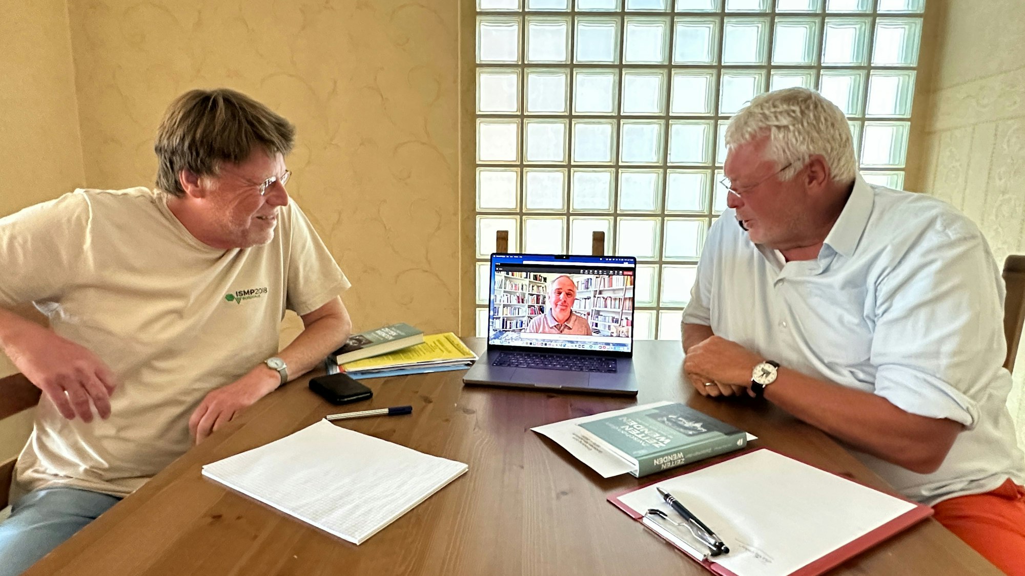 Zwei Männer sitzen an einem Tisch, sie schauen auf ein Tablet, über das ein dritter Mann zugeschaltet ist.