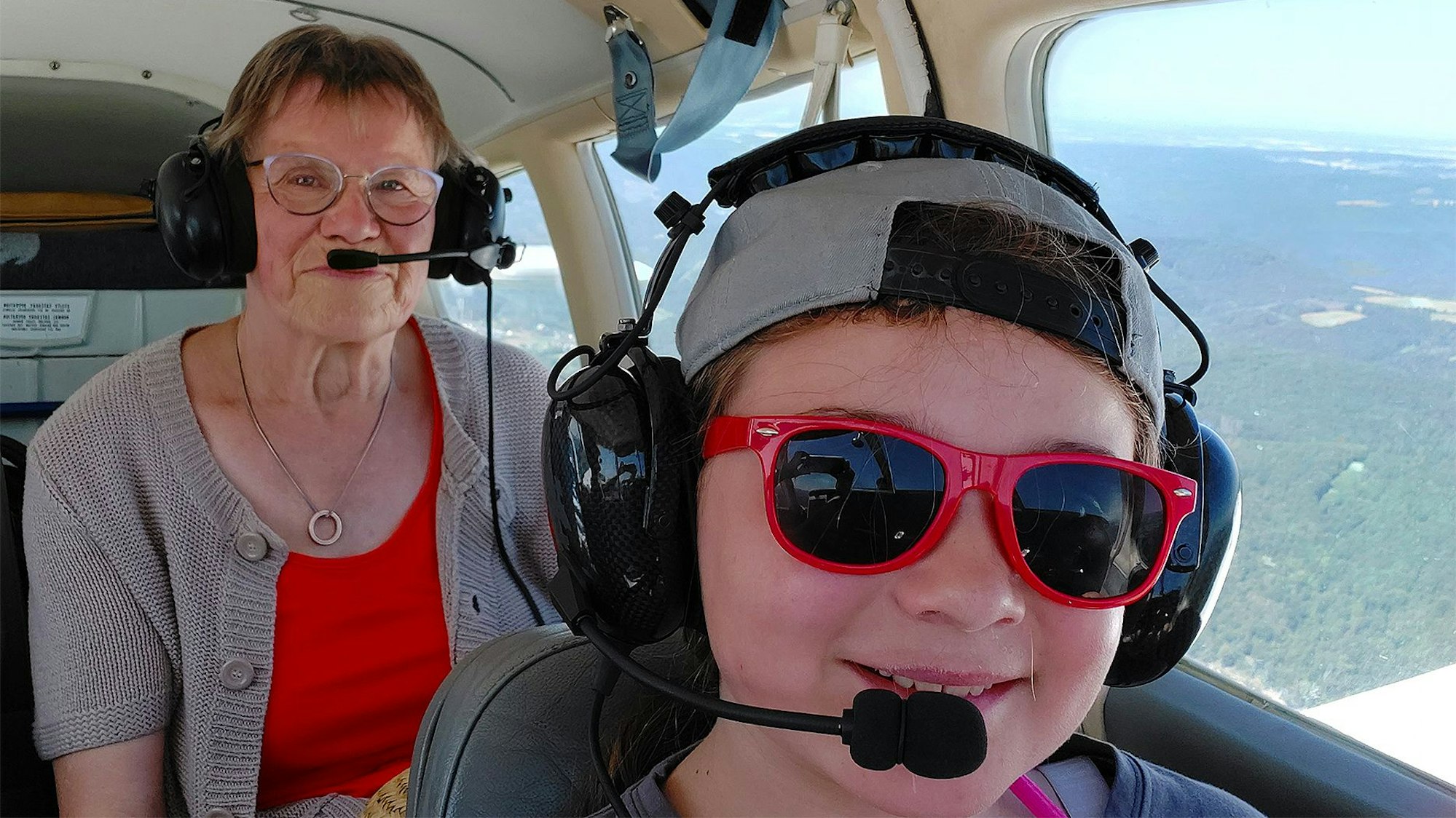Das Bild zeigt ein Kind mit einer roten Sonnenbrille und eine Frau in einem kleinen Flugzeug. Beide tragen Kopfhörer mit Mikrofon.