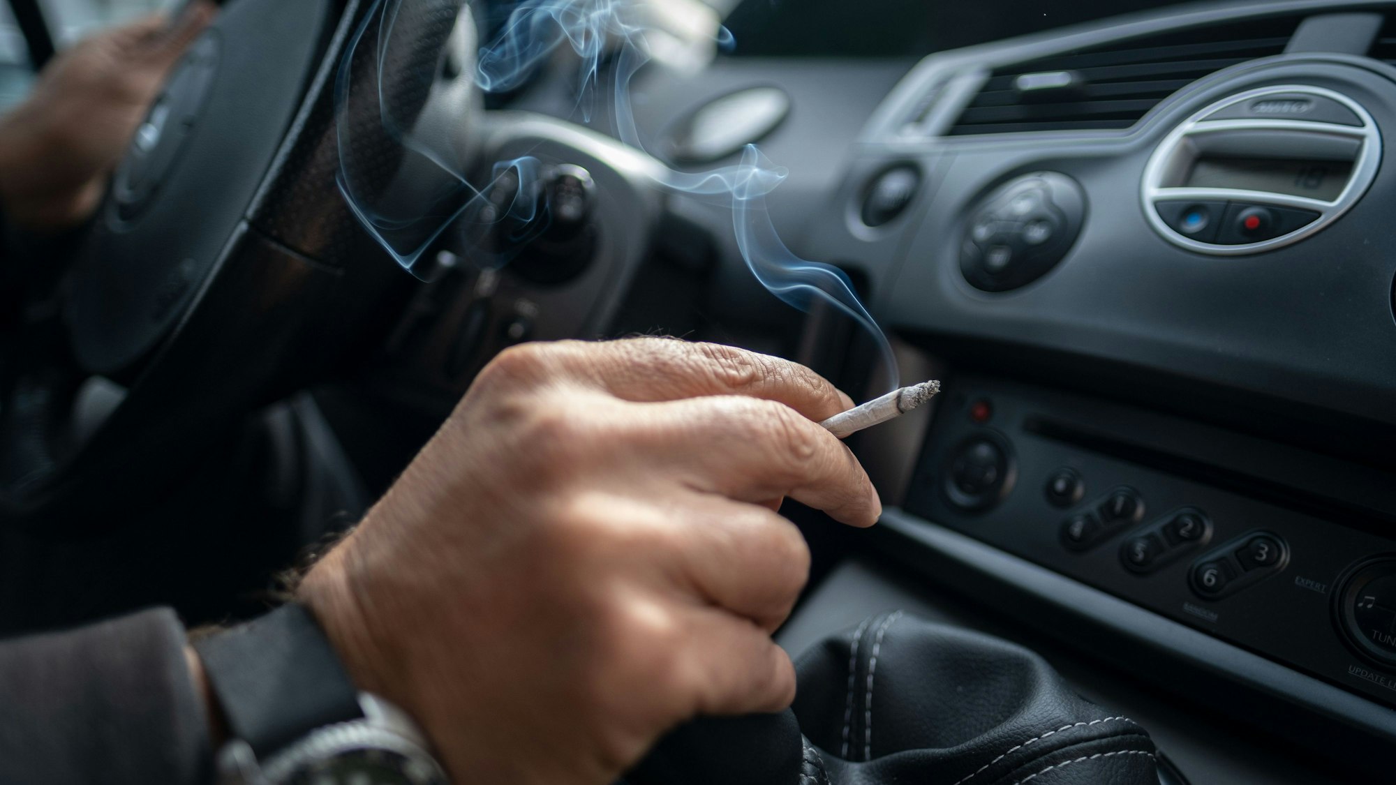 Ein Mann sitzt rauchend am Lenkrad eines Autos.