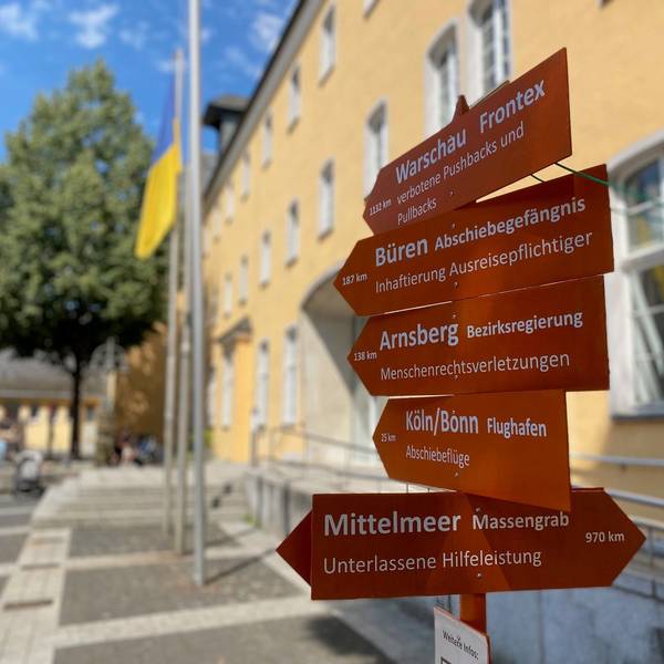 Die hölzernen Wegweiser tragen die Inschrift Mittelmeer, Massengrab und Flughafen Köln/Bonn Abschiebeflüge. Sie stehen vor dem Rathaus.