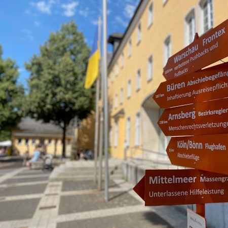 Die hölzernen Wegweiser tragen die Inschrift Mittelmeer, Massengrab und Flughafen Köln/Bonn Abschiebeflüge. Sie stehen vor dem Rathaus.