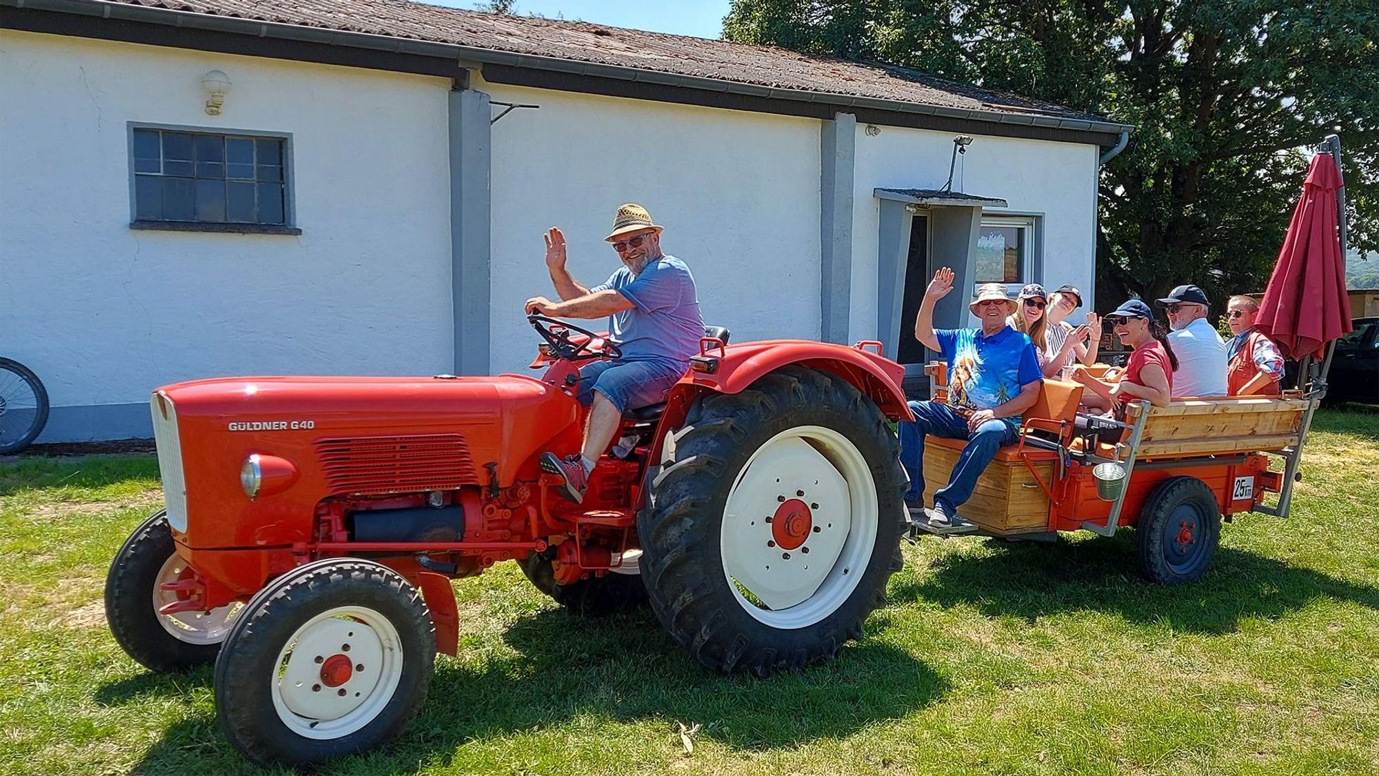 Das Bild zeigt einen winkenden Mann auf einem roten Oldtimer-Traktor. Im Anhänger dahinter sitzt eine ebenfalls winkende Gruppe Menschen.