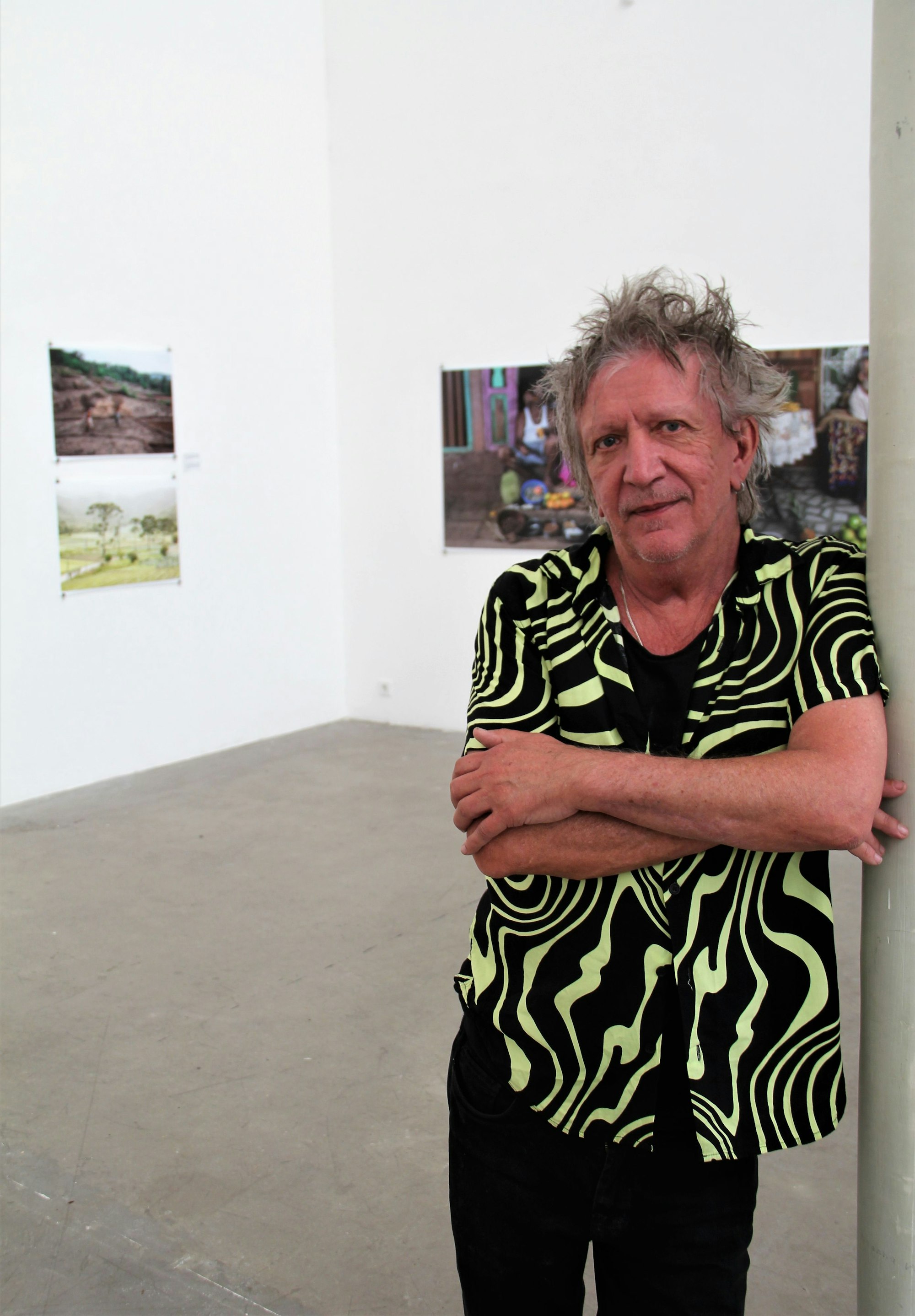 Ein Man mit verstrubbelten grauen Haaren steht mit verschränkten Armen in einem Ausstellungsraum, in dem seine Bilder hängen.