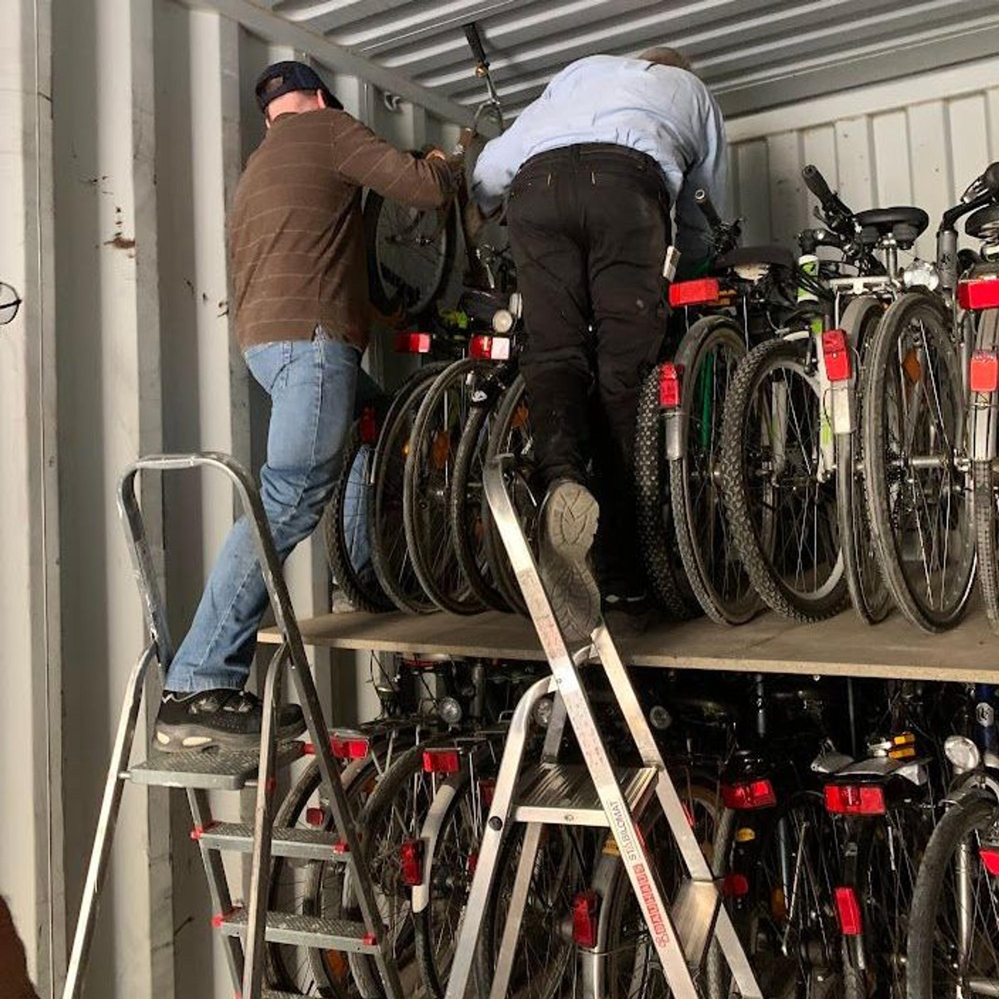 Zwei Männer stehen auf Leitern und stapeln Fahrräder in einem Container übereinander.