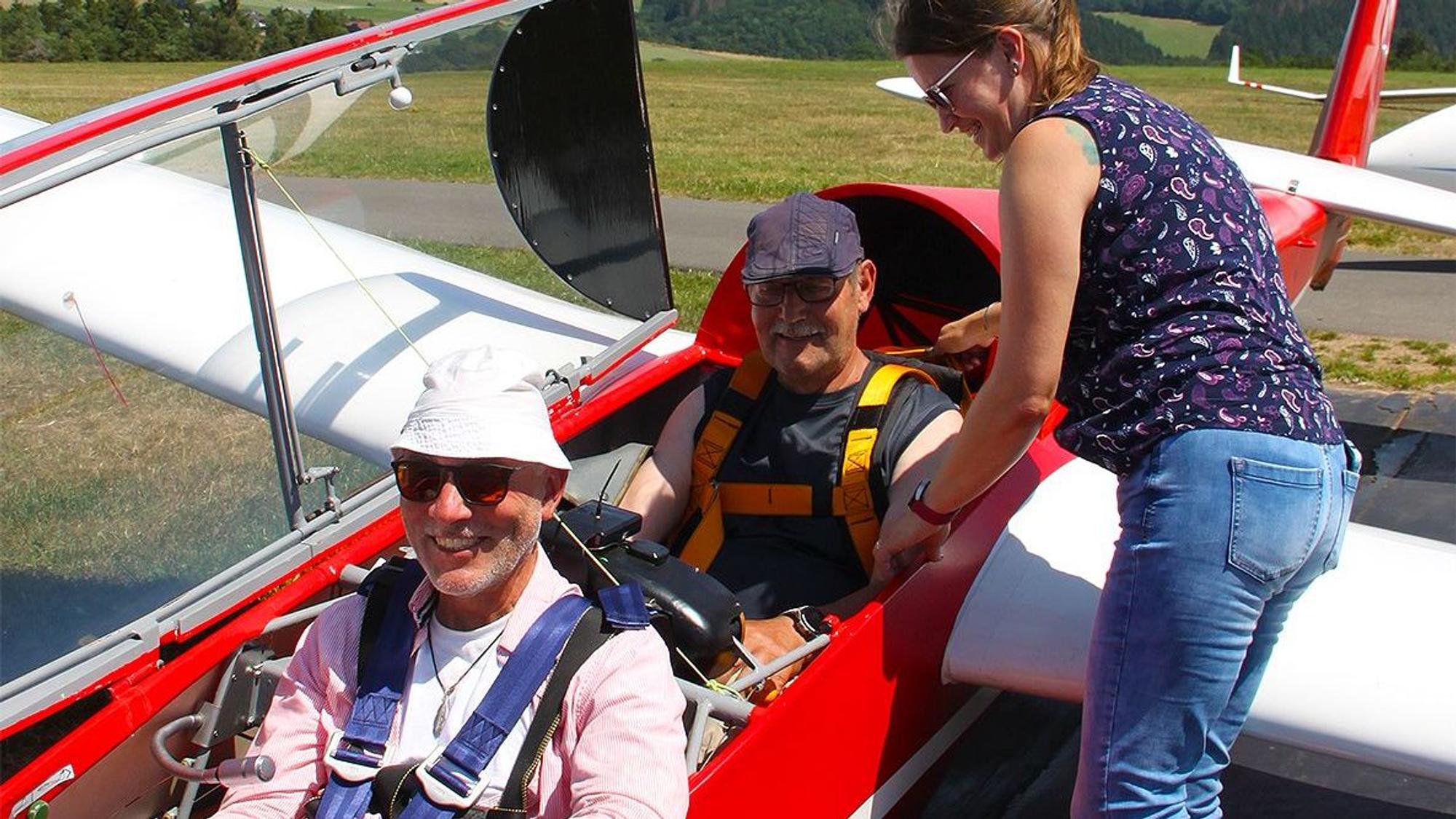 Das Bild zeigt einen Blick in ein Segelflugzeug kurz vor dem Start: Der Pilot sitzt vorne, der Passagier dahinter. Eine Frau hilft ihm beim Anlegen der Sicherheitsgurte.