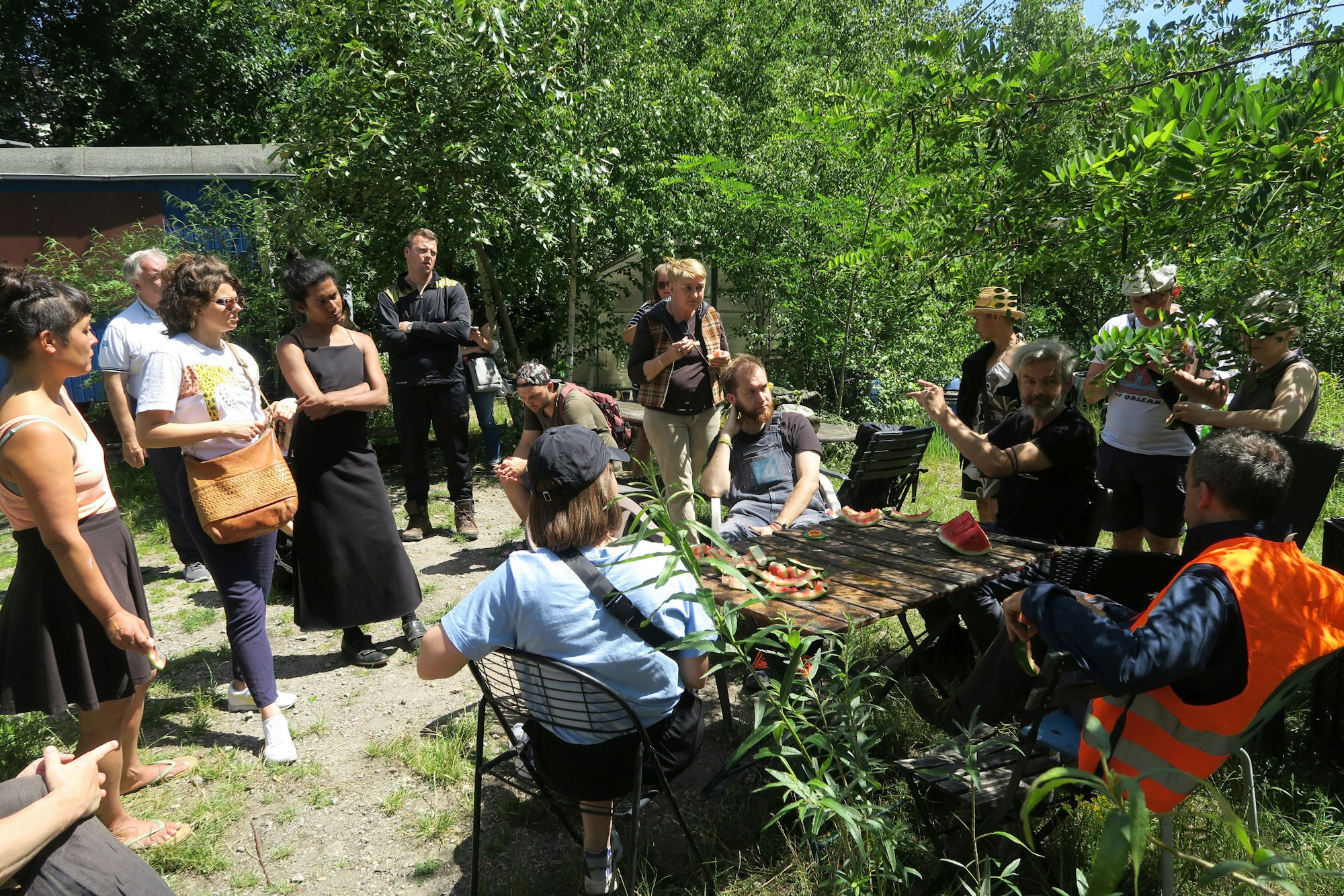 Viele Menschen stehen um einen Tisch im Grünen, einige stehen daneben.