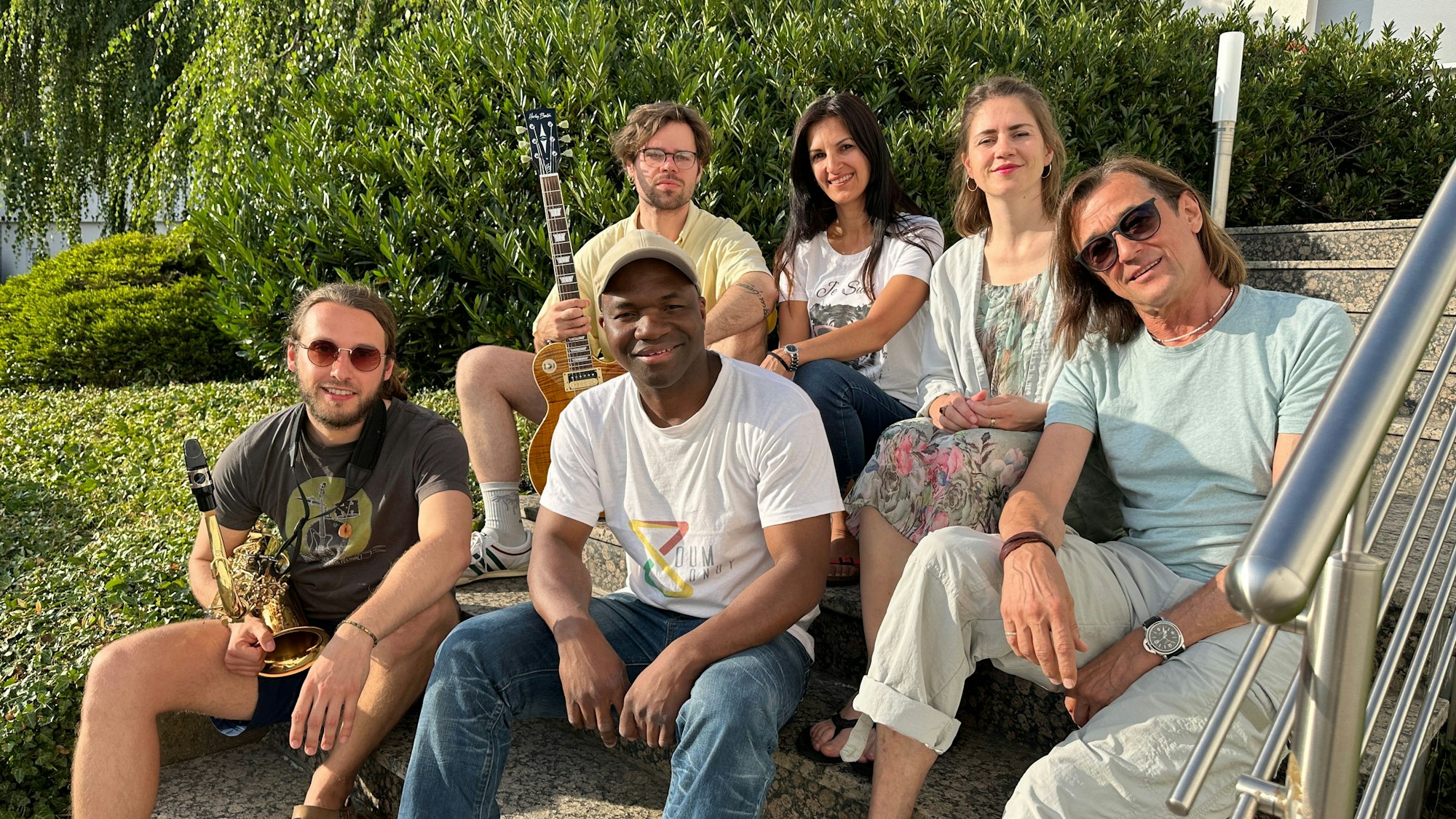 Sechs junge Leute sitzen zum Teil mit Instrumenten in der Hand auf einer breiten Treppe im Freien.