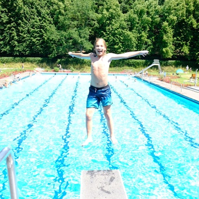 Ein Junge springt rückwärts vom Drei-Meter-Brett, im Hintergrund ist das Schwimmbecken zu sehen.