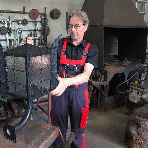 Das Bild zeigt Schmied Albert Köller in seiner Werkstatt. Er hat eine große Lampe in der Hand.