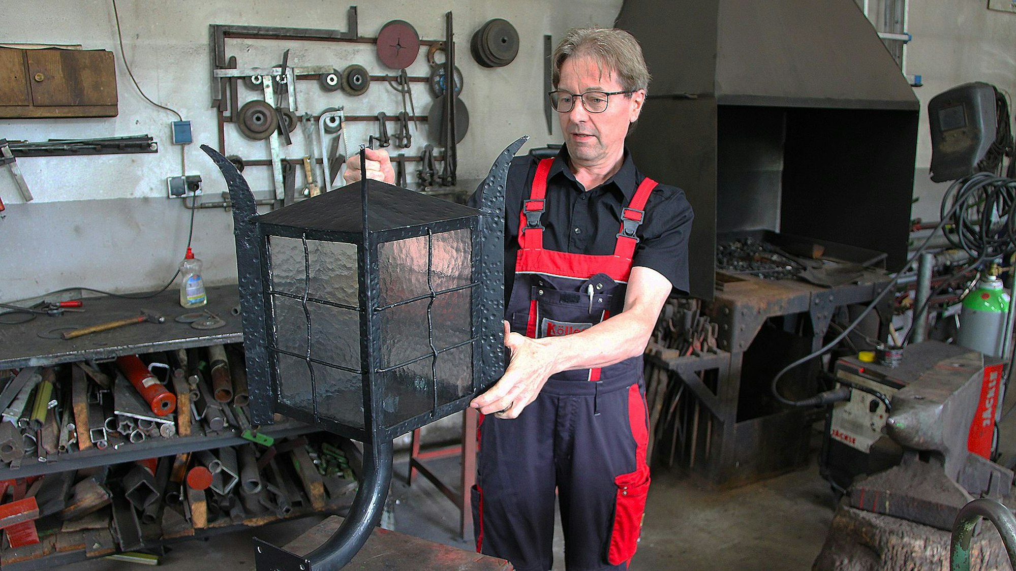 Das Bild zeigt Schmied Albert Köller in seiner Werkstatt. Er hat eine große Lampe in der Hand.